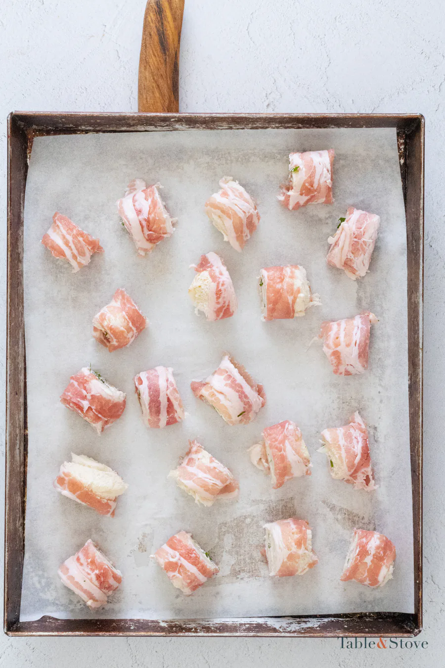 Twenty unbaked bacon-wrapped bread bites spaced across a lined baking sheet