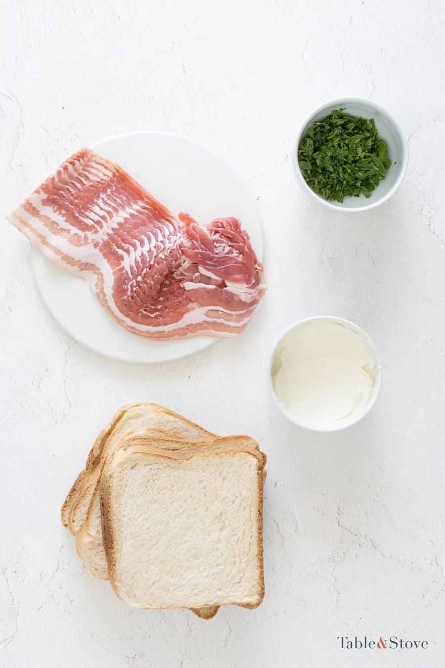Bread, bacon, cream cheese, and chopped herbs arranged on a pale surface