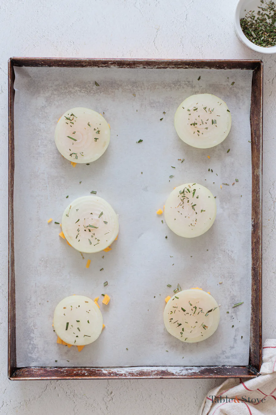 Onion rounds and chopped rosemary arranged over cheddar portions on a baking sheet