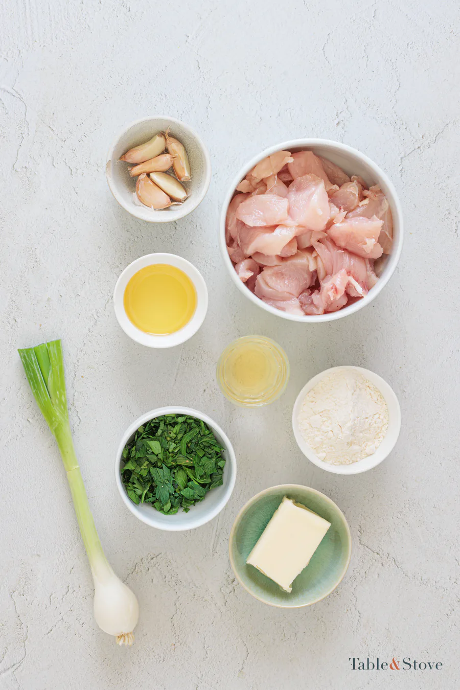 Chicken, flour, garlic, butter, oil, wine, green onion, and parsley arranged on a pale surface