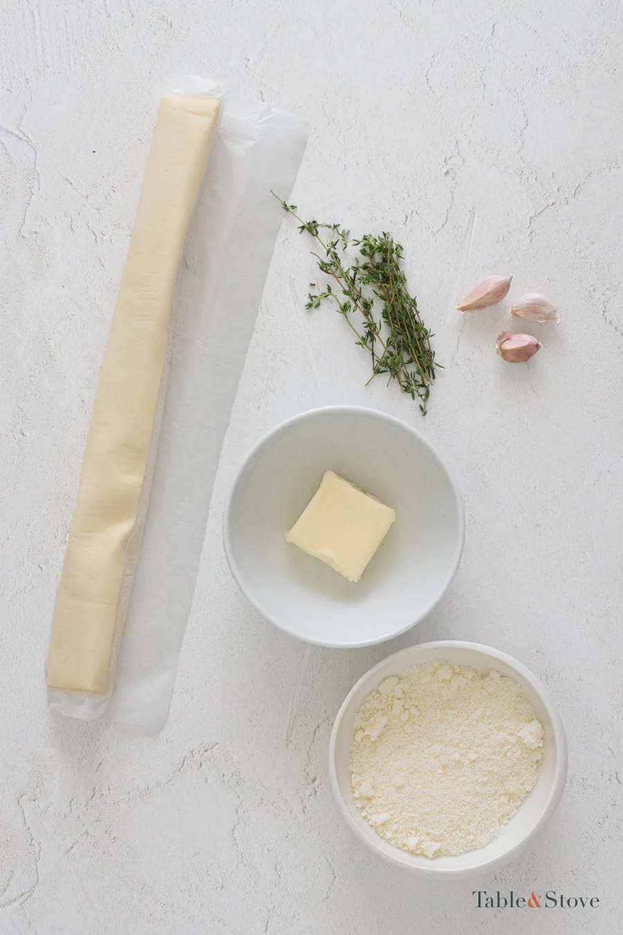 Puff pastry, butter, garlic, Parmesan, and thyme arranged on a pale surface