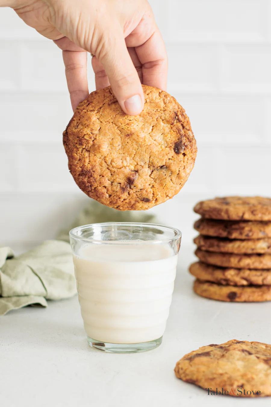 Hand holding a baked oat cookie above a glass of milk