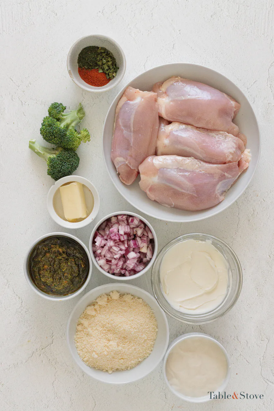 Chicken thighs, broccoli, cheeses, cream, butter, onion, salsa verde, and seasonings arranged on a pale surface