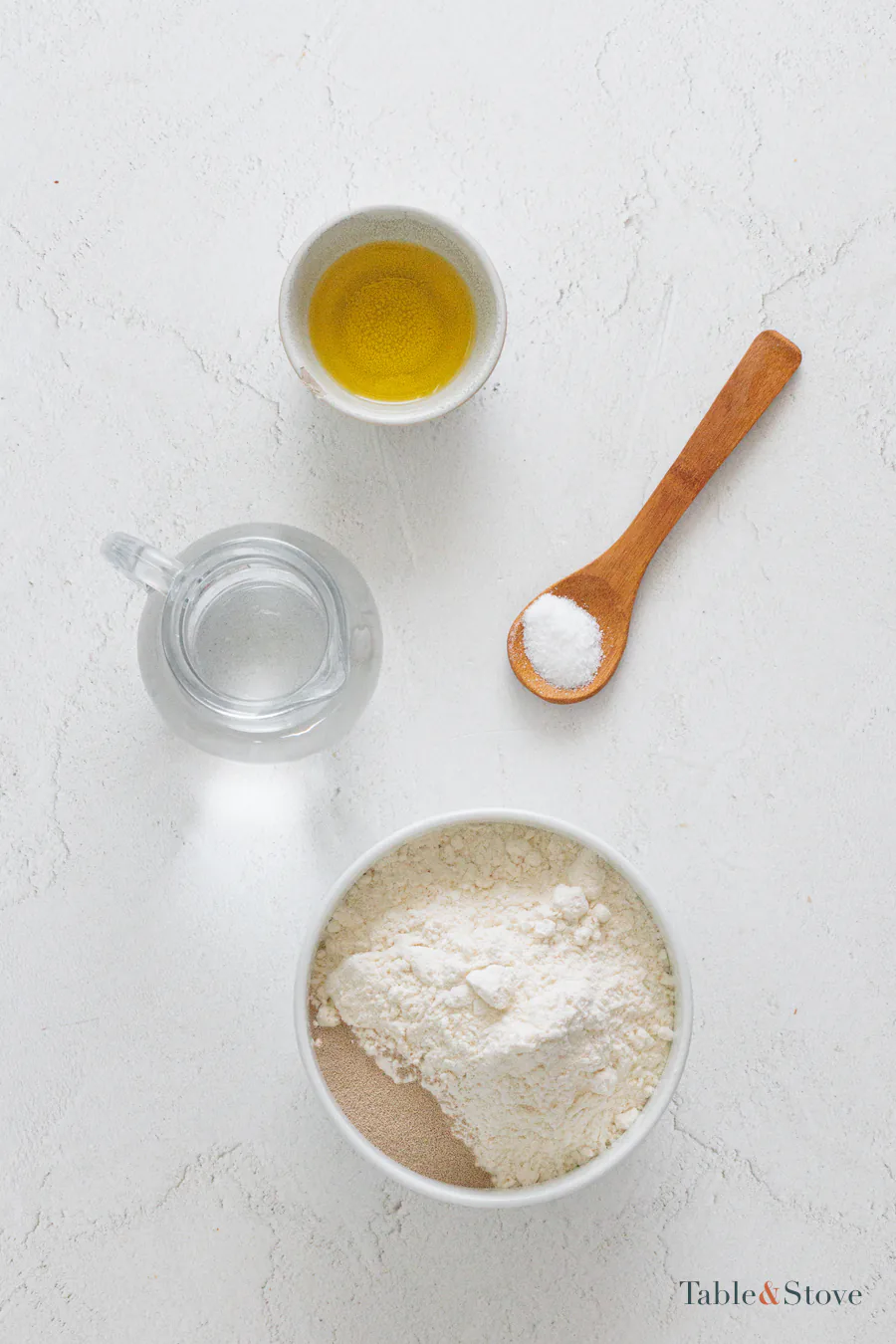 Flour, water, olive oil, salt, and dry yeast arranged on a pale surface