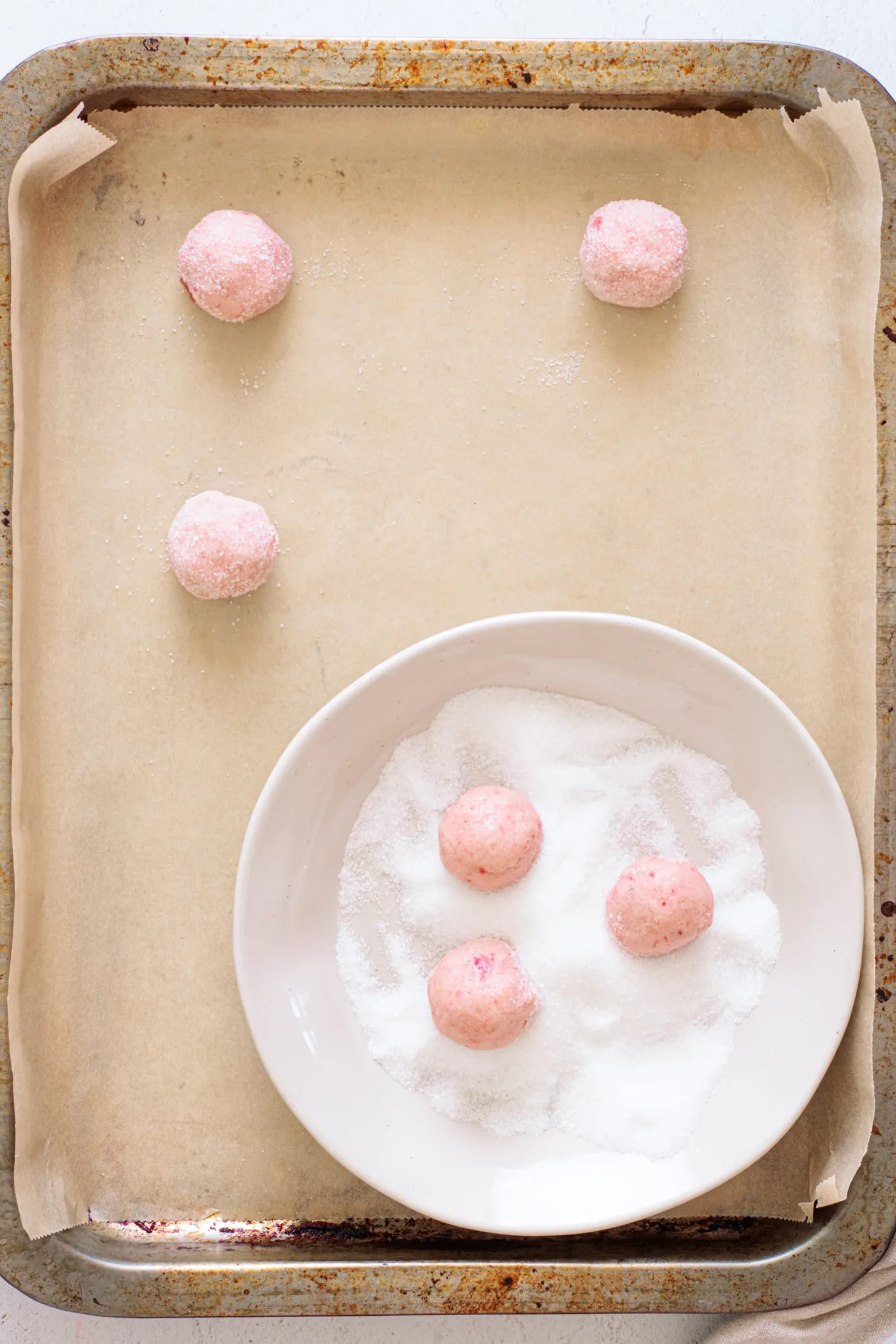 Strawberry cookie dough balls being rolled in granulated sugar beside a lined baking sheet